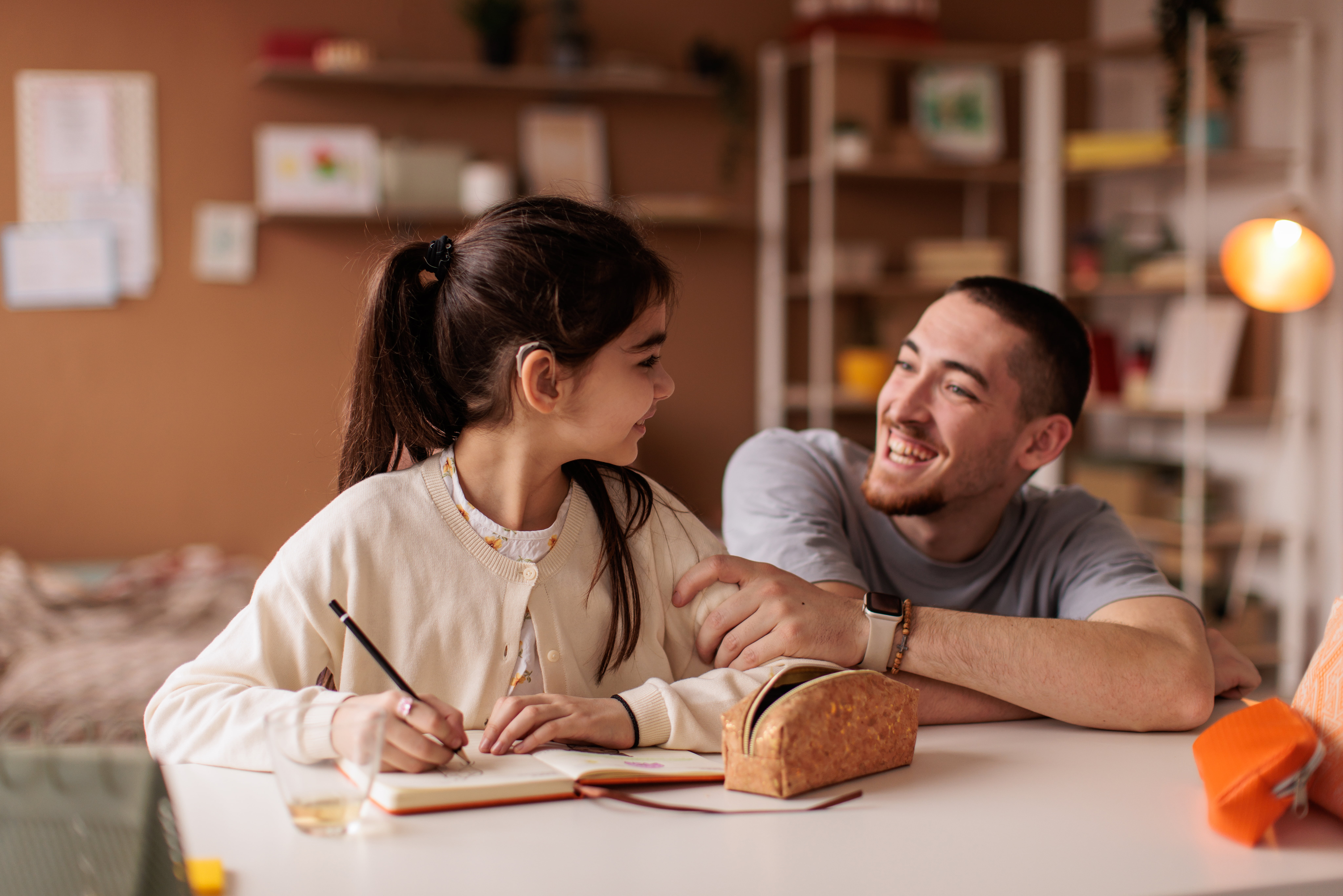 Jente med høreapparat tegner i en bok ved et bord i stuen sammen med en mann.