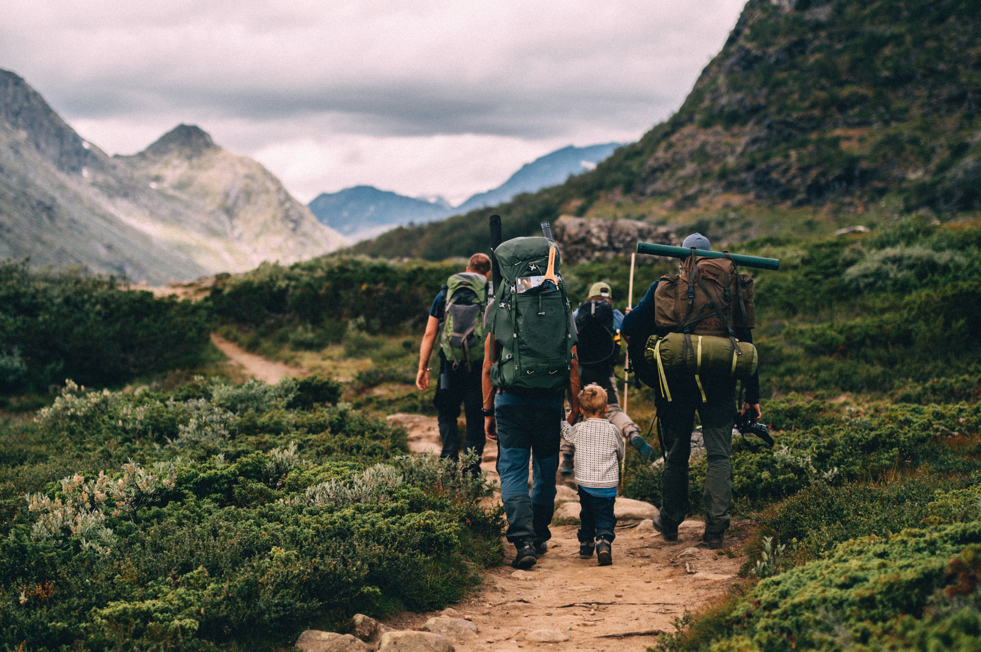 Gruppe mennesker med ryggsekker og campingutstyr går på tursti i fjellandskap med grønn vegetasjon og høye fjell under overskyet himmel.
