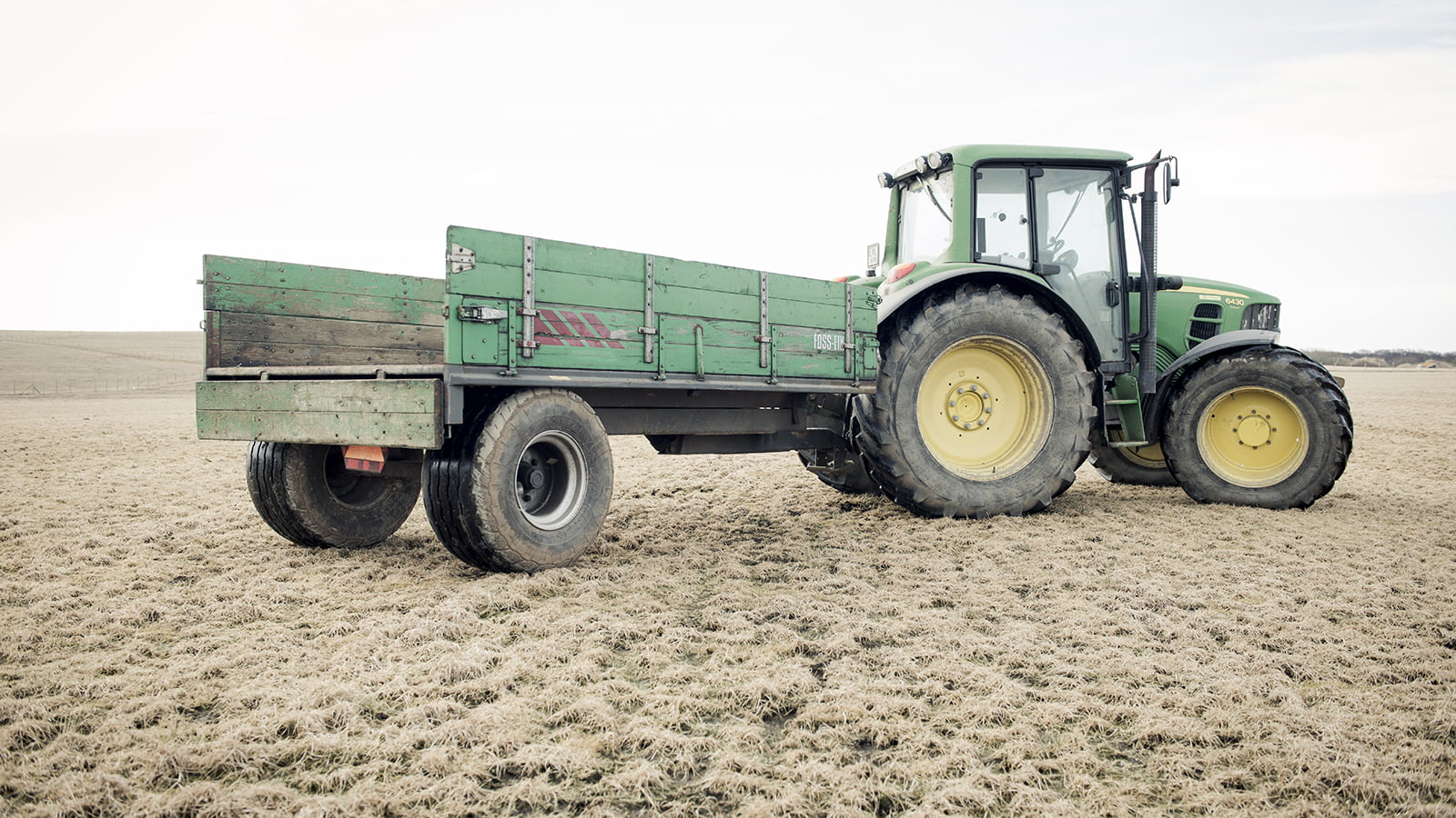 Grønn traktor med tilhenger står på et åpent jorde under en lys himmel.